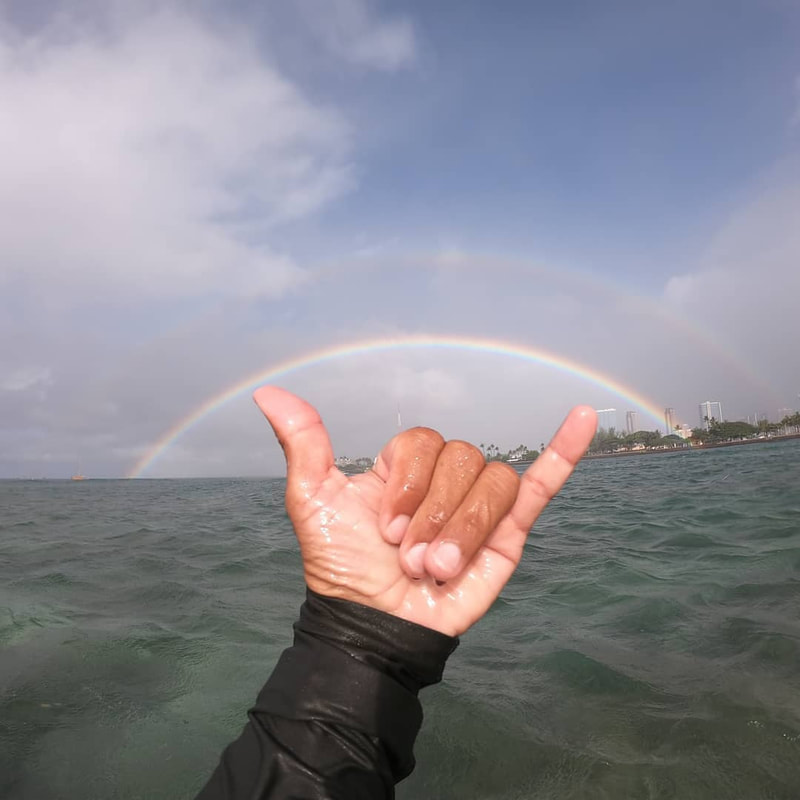 A shaka with a beautiful double rainbow above it over the water. Provided by Polu Lani Surf.